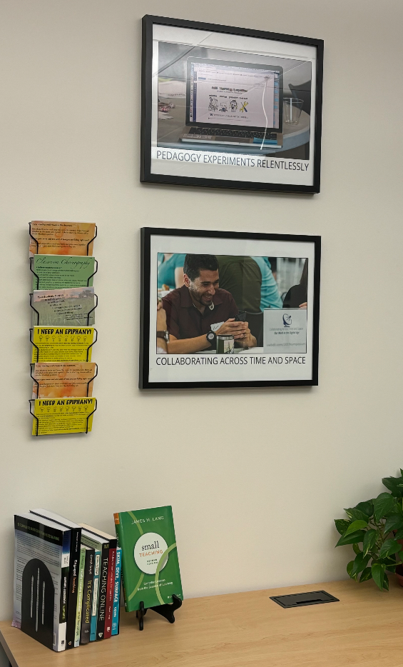 Office pace with a table and chairs. On the wall are a couple of framed prints and a card rack. The table has some books about teaching and a plant. 